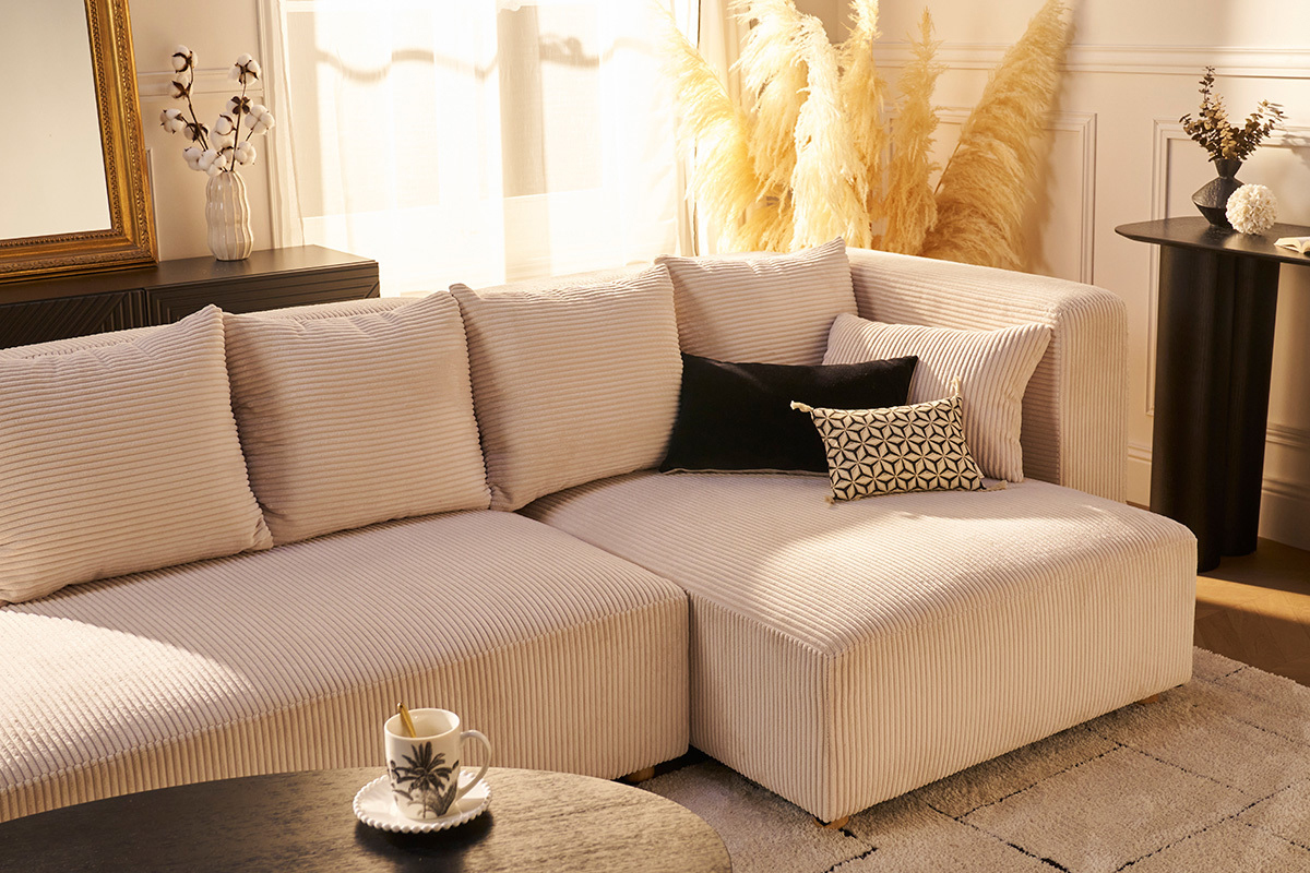 Canap� d'angle droit 3 places en velours c�tel� beige avec coussins dans un salon �clair�.