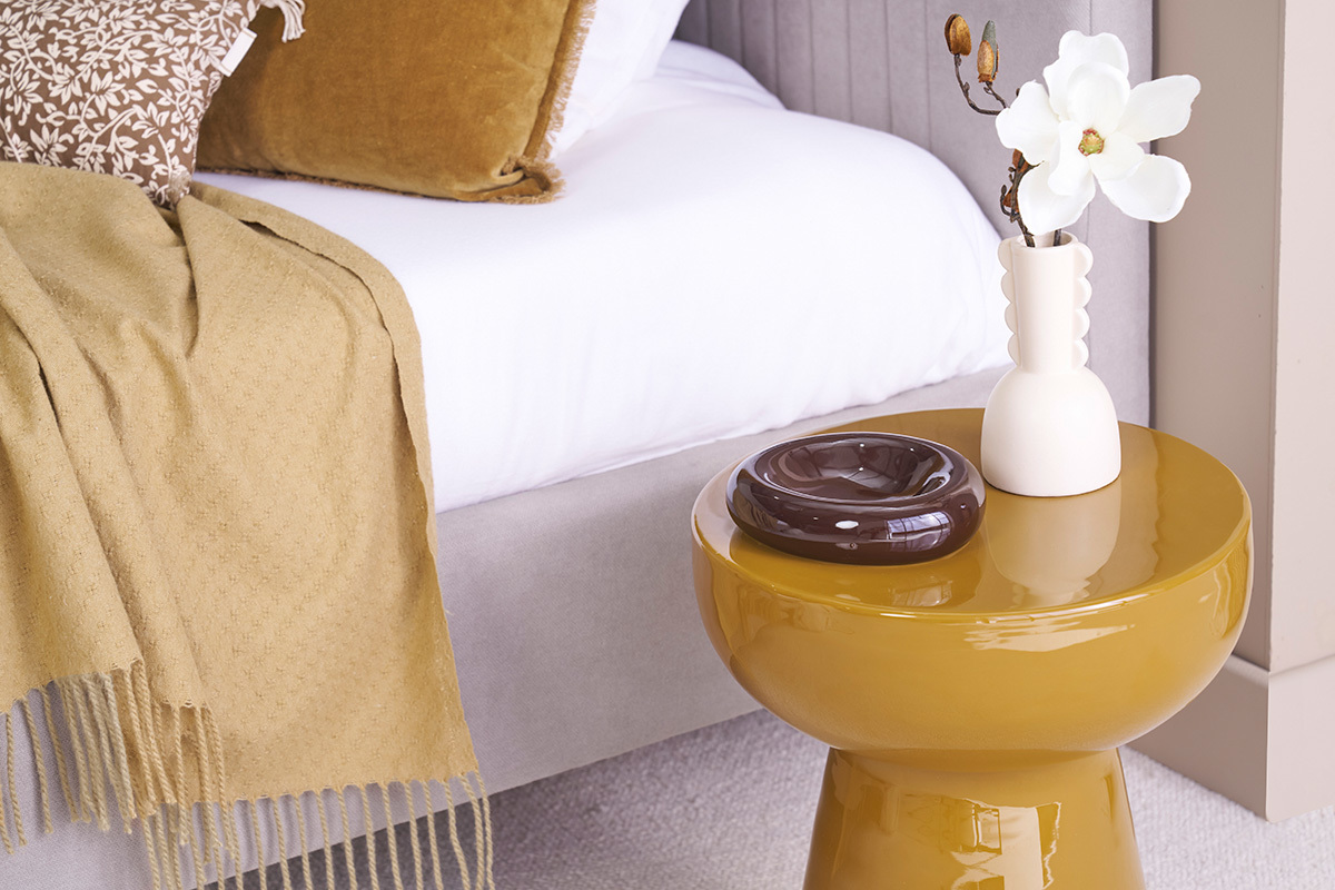 Table d'appoint MOGOU ronde bronze avec vase blanc et cendrier marron, � c�t� d'un lit.